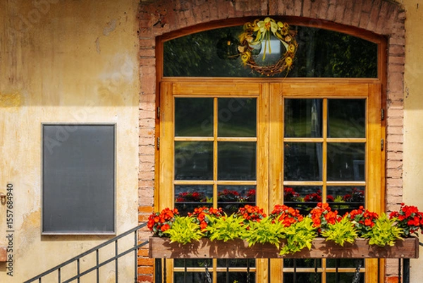 Fototapeta Charming restaurant facade with wooden-framed windows, red flowers in a planter, a decorative wreath.