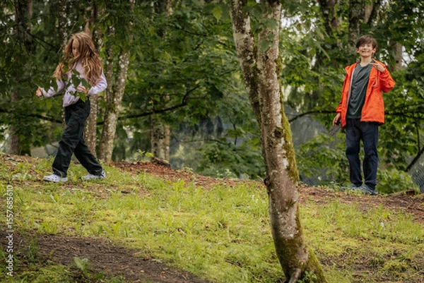 Fototapeta Two children explore a forested park, with one walking and the other smiling at the camera. The ground is grassy and slightly sloped with tree cover.