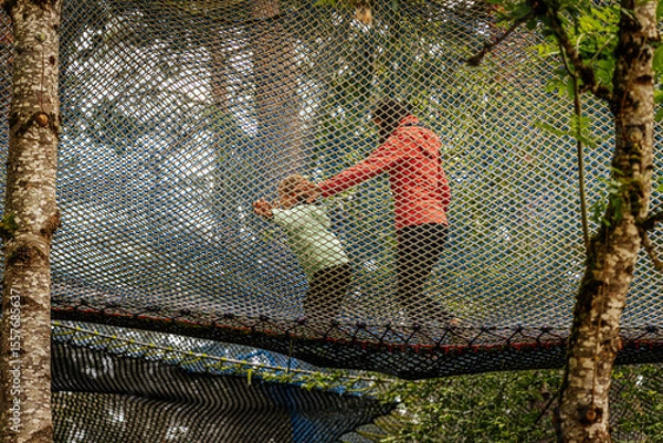Fototapeta An adult and child walk together on a large safety net suspended among trees in a forest adventure park, creating a fun and active bonding moment.