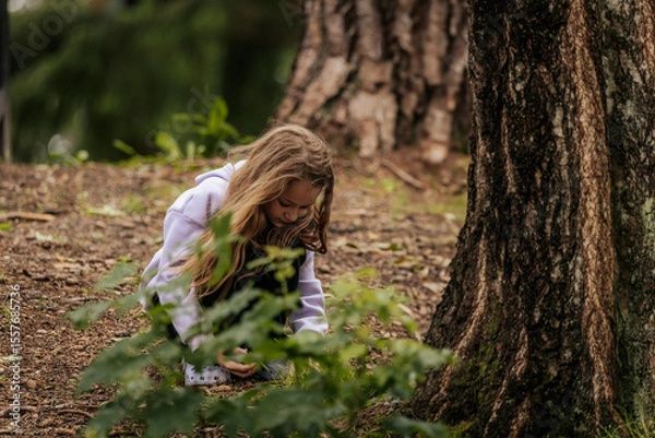 Fototapeta A young girl kneels on a forest floor, curiously examining something in the soil near a large tree trunk, surrounded by greenery and natural textures.