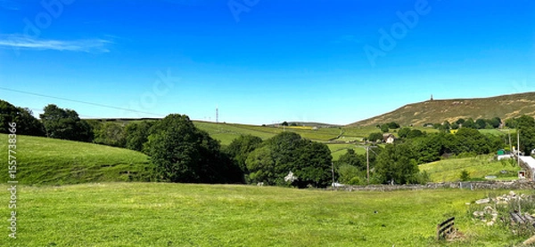 Fototapeta Rolling green hills stretch across the landscape under a clear blue sky. A few scattered trees and distant structures add to the serene countryside view in, Lumbutts, Todmorden, UK