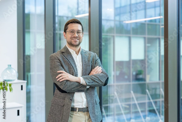 Fototapeta A smiling young man, founder and owner of a firm and company, stands in the lobby of an office building, arms crossed on his chest and confidently looking into the camera