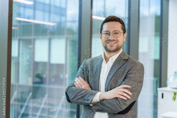 Fototapeta Portrait of a smiling young businessman standing in the office by the window in a suit and glasses, arms crossed on his chest and looking confidently at the camera