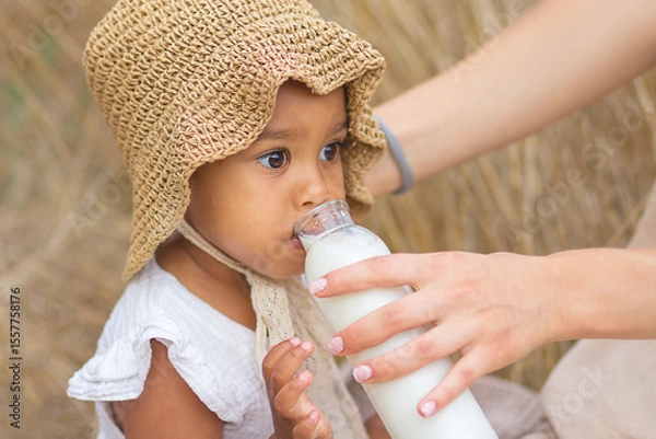 Obraz A large portrait of a little mulatto girl who drinks milk from a bottle held by her mother's hands at a picnic in the field. Healthy eating