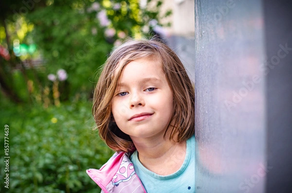 Fototapeta Playful preschooler peeks around pillar with mischievous gaze. Blonde bob, gray-blue eyes, light blue top. Soft summer greenery bokeh. Childhood curiosity close-up