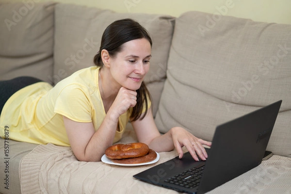Obraz European woman lies on sofa with a laptop and a pastry, enjoying a relaxed moment at home in casual clothing, looking at the screen with a smile