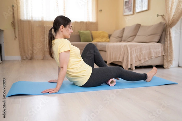 Obraz European woman uses foam roller on mat for back massage and recovery exercise, relaxing in cozy home interior. Gymnastics, fitness, aerobics at home