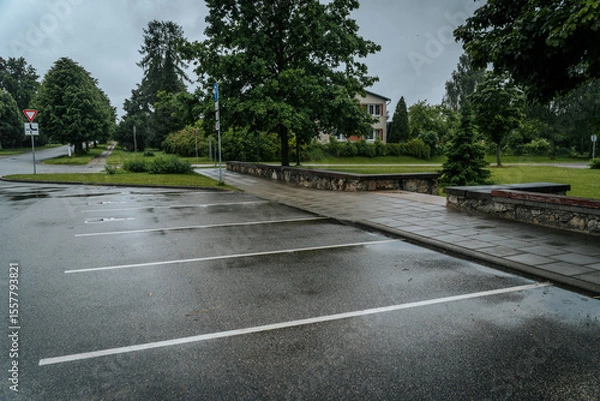 Fototapeta An empty, wet parking lot on a rainy day, with sidewalks, green trees, traffic signs, and a residential building in the background. Overcast sky and copy space.