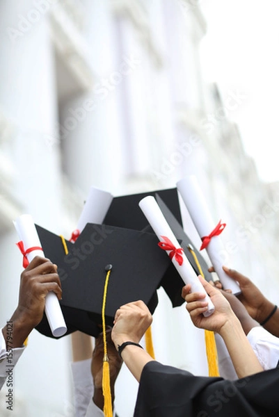 Fototapeta A group of multietnic students celebrating their graduation by throwing caps in the air closeup. Education, qualification and gown concept.