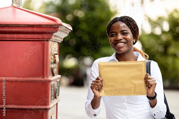 Obraz Portrait of smiling black female tourist standing next to mailbox and holding letter.