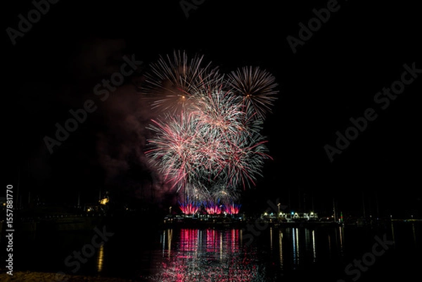 Fototapeta Colorful fireworks exploding over a marina at night, reflected in the calm harbor water with yachts and masts silhouetted under a starless sky with copy space