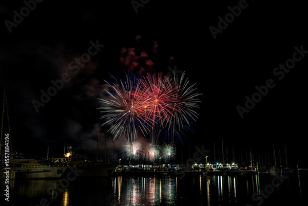 Fototapeta Colorful fireworks exploding over a marina at night, reflected in the calm harbor water with yachts and masts silhouetted under a starless sky with copy space