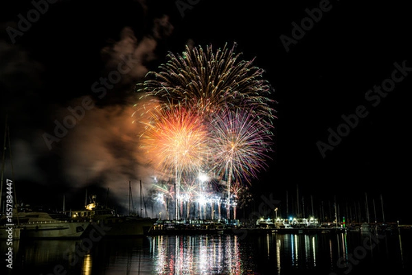 Fototapeta Colorful fireworks exploding over a marina at night, reflected in the calm harbor water with yachts and masts silhouetted under a starless sky with copy space