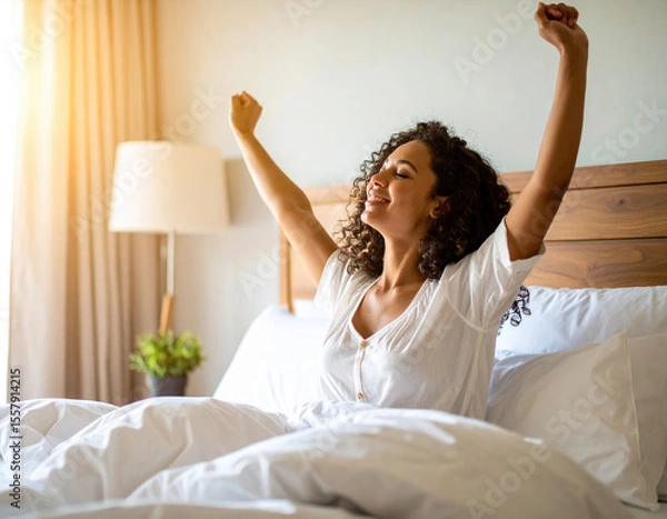 Fototapeta Beautiful woman waking up in cozy bedroom, stretching her arms with joyful expression. soft morning light illuminates room, creating warm and inviting atmosphere