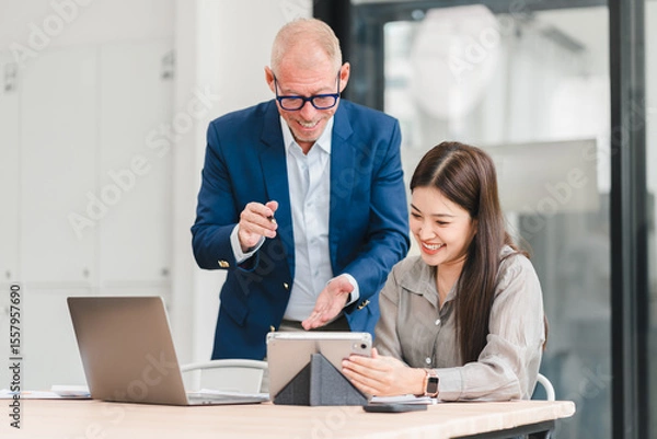 Fototapeta Two business colleagues smiling and discussing work using tablet and laptop in modern office, showing collaboration and positive teamwork atmosphere
