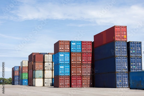 Obraz Colorful stacked shipping containers are neatly arranged under a clear blue sky at a freight yard. The image highlights global trade, logistics, and industrial transportation infrastructure.