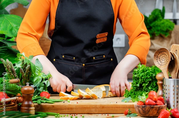 Fototapeta Chef woman in a black apron skillfully slices baguette bread on a cutting board surrounded by fresh greens and healthy ingredients in a cozy kitchen.