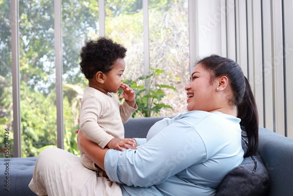 Obraz Mother and little son are sitting and relaxing on the sofa at home. They are smiling happily. Family concept. Learning development for 1 year old children.