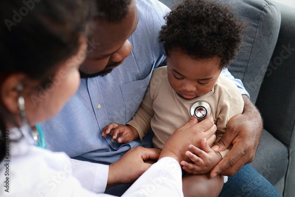 Obraz Doctor uses stethoscope to examine heart of little boy sitting on father's lap to check for possible abnormalities in child. Child health concept. Medical services.
