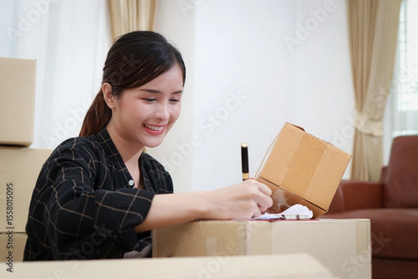 Obraz Beautiful Asian female entrepreneur doing online business, writing product information on a parcel box to send to customers. Internet service. Online business idea.