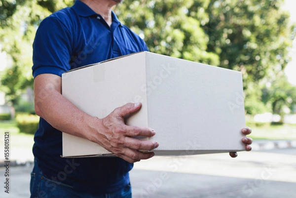 Obraz Delivery man wearing blue shirt working in delivery service, holding box to deliver to customer at doorstep. Transporting goods, parcels.