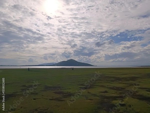 Fototapeta Scenic wide-angle shot of a lush green field by a calm lake, with a mountain in the distance under a dramatic cloudy sky. A peaceful nature scene capturing open space and atmospheric light.