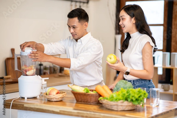 Obraz Asian man using blender making smoothie drink while woman holding apple watching at kitchen counter