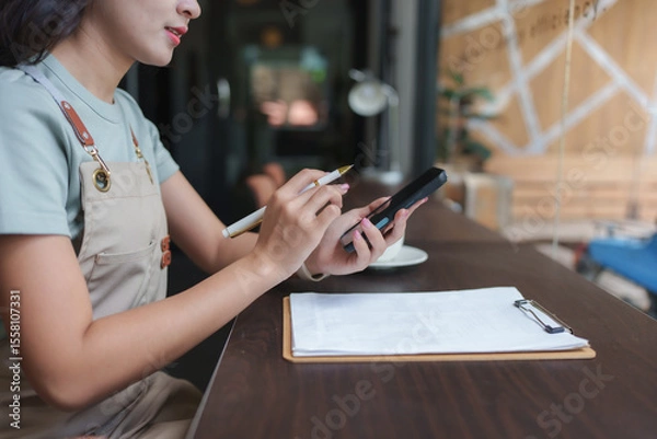 Obraz Coffee shop owner managing inventory and orders using smartphone and clipboard