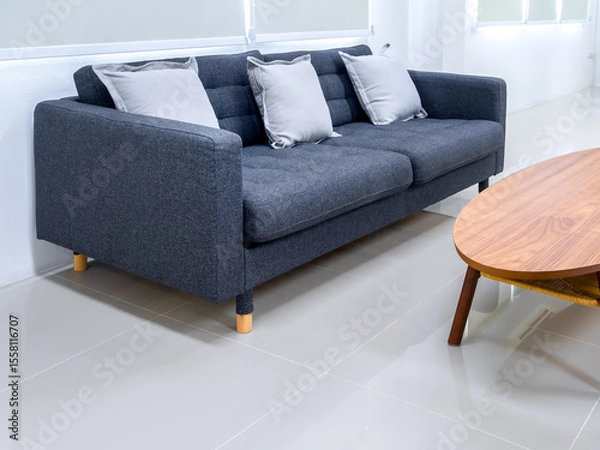 Obraz Empty grey cozy fabric sofa seat with pillows near oval wooden table on clean tiles floor, near roller blinds at the window, wait area on white modern living room with nobody.