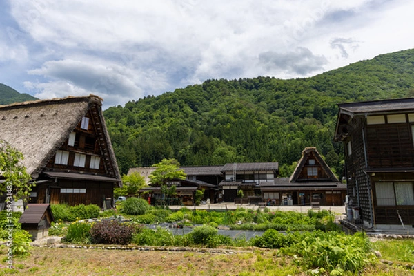 Fototapeta 白川郷の合掌造り家屋と畑、庭園。青々とした山々と初夏の雲が広がる伝統的な美しい風景。
