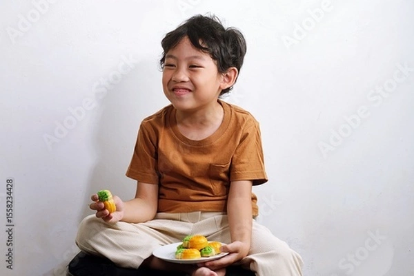 Obraz Happy Asian boy eats traditional nastar pineapple tart at home. A candid and joyful moment of childhood with local Indonesian snack.