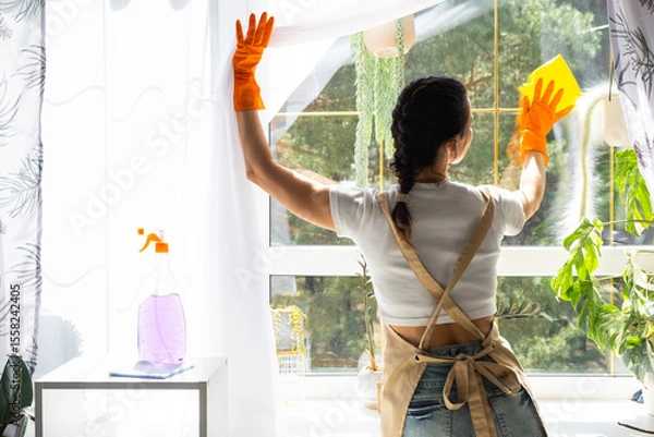 Fototapeta Woman in rubber gloves and apron manually washes window of house with rag cleaner and mop inside interior with home plants on windowsill. Restoring order and cleanliness in spring, cleaning servise