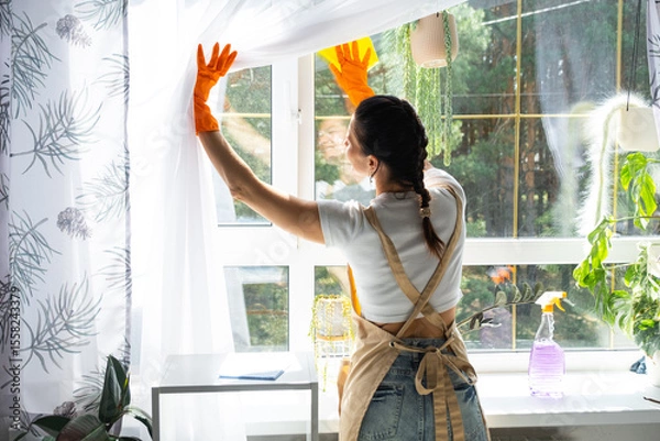 Fototapeta Woman in rubber gloves and apron manually washes window of house with rag cleaner and mop inside interior with home plants on windowsill. Restoring order and cleanliness in spring, cleaning servise