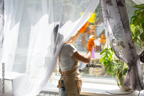 Fototapeta Woman in rubber gloves and apron manually washes window of house with rag cleaner and mop inside interior with home plants on windowsill. Restoring order and cleanliness in spring, cleaning servise