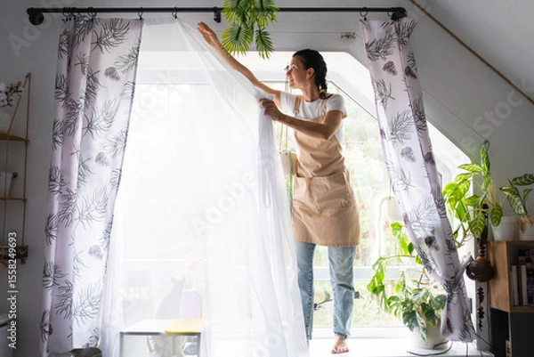 Fototapeta Woman in apron hangs transparent tulle curtains on large attic windows in the house inside the interior with potted plants. Spring cleaning, tidying up
