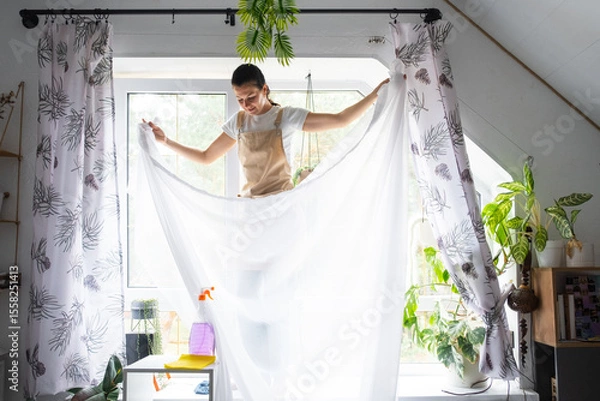 Fototapeta Woman in apron hangs transparent tulle curtains on large attic windows in the house inside the interior with potted plants. Spring cleaning, tidying up