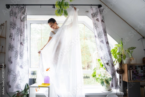 Fototapeta Woman in apron hangs transparent tulle curtains on large attic windows in the house inside the interior with potted plants. Spring cleaning, tidying up