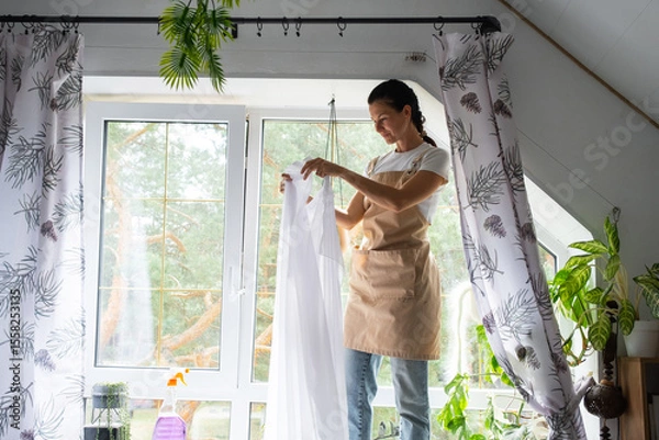 Fototapeta Woman in apron hangs transparent tulle curtains on large attic windows in the house inside the interior with potted plants. Spring cleaning, tidying up