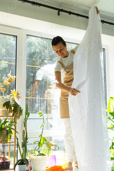 Fototapeta Woman in apron hangs transparent tulle curtains on large windows in the house inside the interior with potted plants. Spring cleaning, tidying up