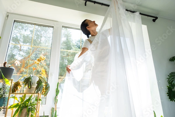 Fototapeta Woman in apron hangs transparent tulle curtains on large windows in the house inside the interior with potted plants. Spring cleaning, tidying up