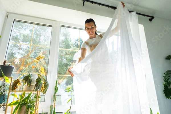Fototapeta Woman in apron hangs transparent tulle curtains on large windows in the house inside the interior with potted plants. Spring cleaning, tidying up