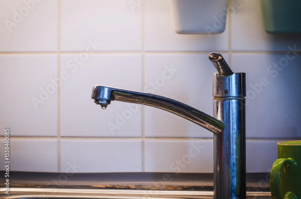 Fototapeta Old chrome kitchen faucet with dripping water drops captured in closeup against a white ceramic tiled wall background showing plumbing detail and water waste concept.