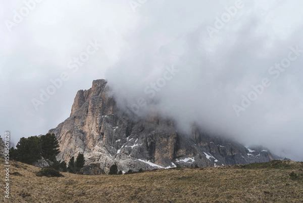 Fototapeta Dramatic view of Sassolungo mountain partly covered by clouds, with rocky cliffs and grassy alpine meadow in the Dolomites, Italy.