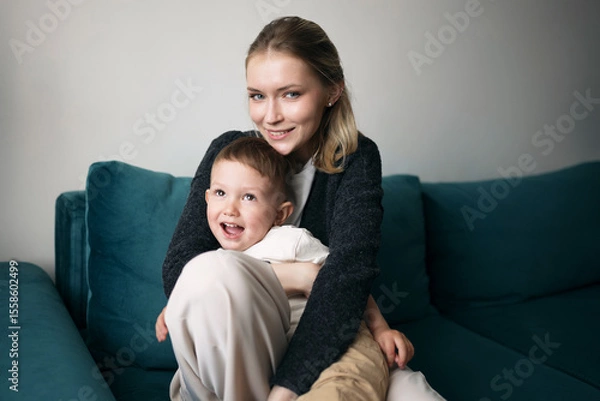 Fototapeta Smiling young mother hugging her cheerful son while sitting on a modern teal sofa at home, capturing a moment of joy and family love.