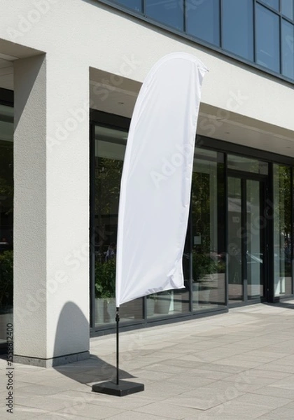 Fototapeta A mockup of a tall, blank white feather flag standing outside a modern storefront on a sunny day. The flag is gently waving in the breeze. The background shows a clean, modern building facade