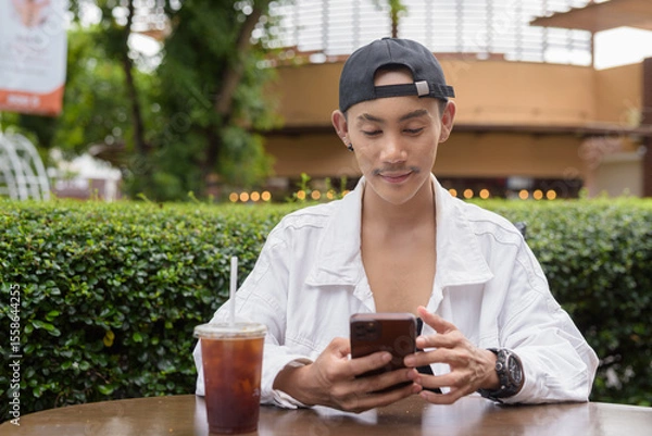 Obraz Non binary gay man drinking iced coffee in outdoors cafe restaurant and using mobile phone