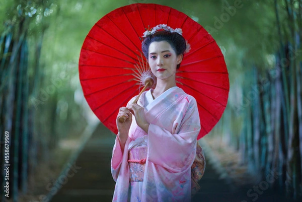 Fototapeta A young Asian Japanese woman in a Traditional Furisode Kimono Yukata dress costume holding a red wagasa umbrella in a natural bamboo tunnel.  An old-fashioned cultural dress of a Japanese girl.