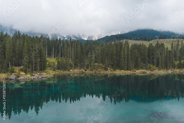 Fototapeta Lake Carezza in Dolomites, Italy, with clear turquoise water, pine forest, and misty mountains in the background.