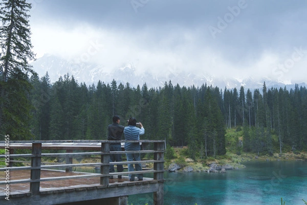 Fototapeta Couple standing on wooden terrace enjoying mountain lake view with forest and cloudy sky, peaceful nature moment.
