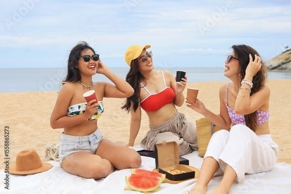 Obraz Three happy young Asian women enjoying pizza and drinks during a beach picnic on a sunny summer day. Group of friends relaxing on vacation, laughing and having fun outdoors near the sea.